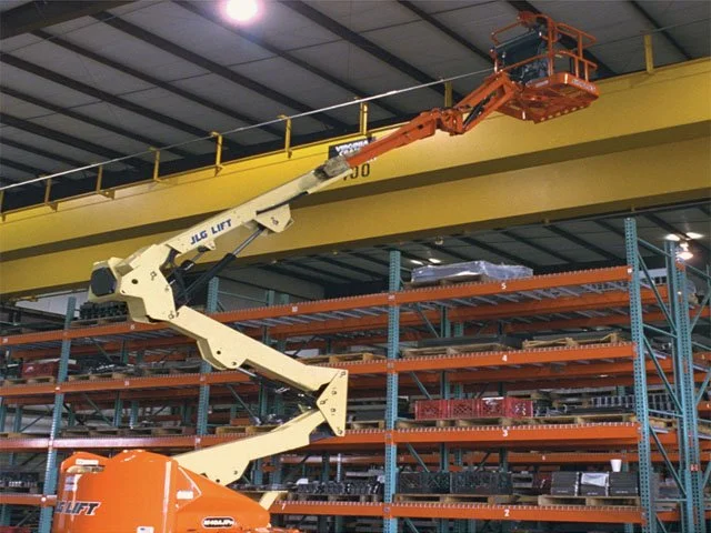 Grúa articulada en un almacén, elevando una viga amarilla.