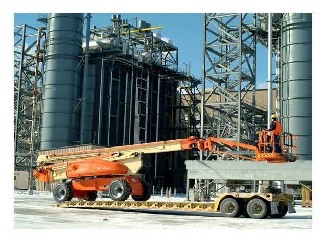 Una torre industrial y maquinaria de construcción con un trabajador usando un elevador en una estructura metálica.
