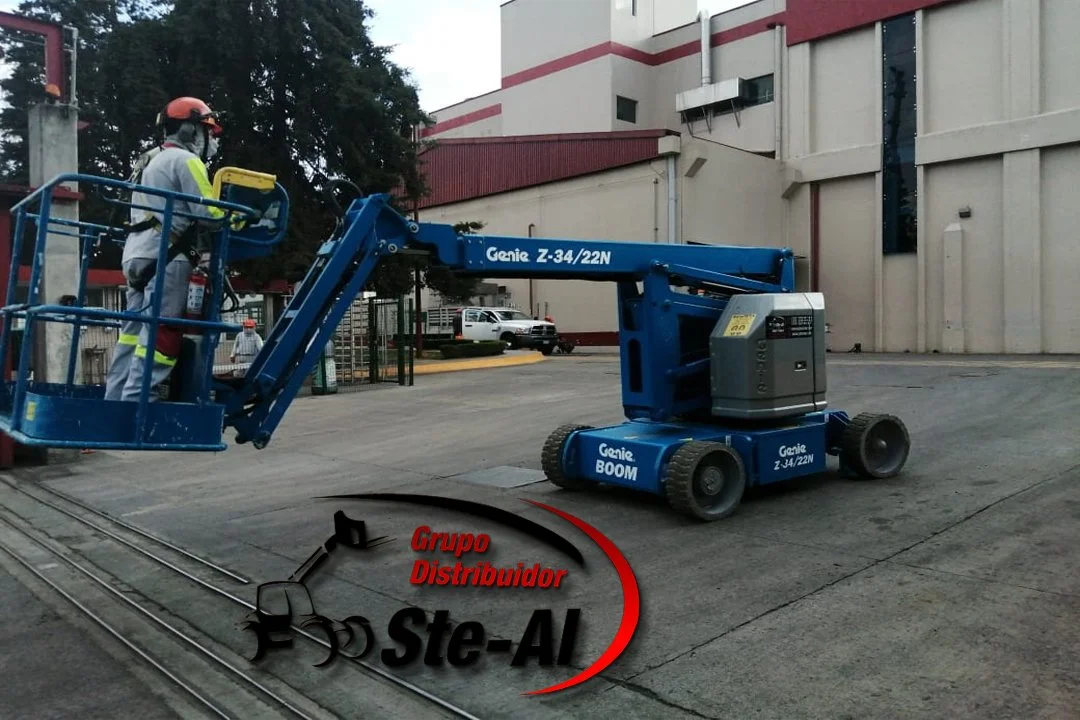 Persona operando un elevador de plataforma azul en un estacionamiento frente a un edificio con paredes blancas y detalles en rojo.